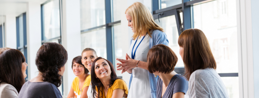 A group of women seated around a table in a bright meeting room, listening to a standing woman leading the discussion.